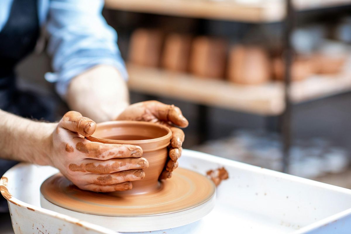 陶匠雙手在轆轤上塑造陶器，身後架上是成品 · a potter's hands shaping a vessel on the wheel, finished pots racked behind